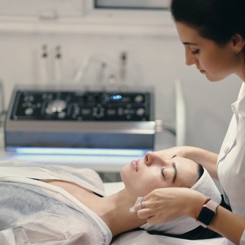 Woman lying during cosmetic procedure while skillful female cosmetologist making facial treatment