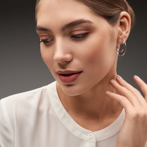 Close up portrait of young beautiful brunette female model presenting silver earrings. Front view of woman with clean skin posing, isolated on studio background. Concept of jewelry.